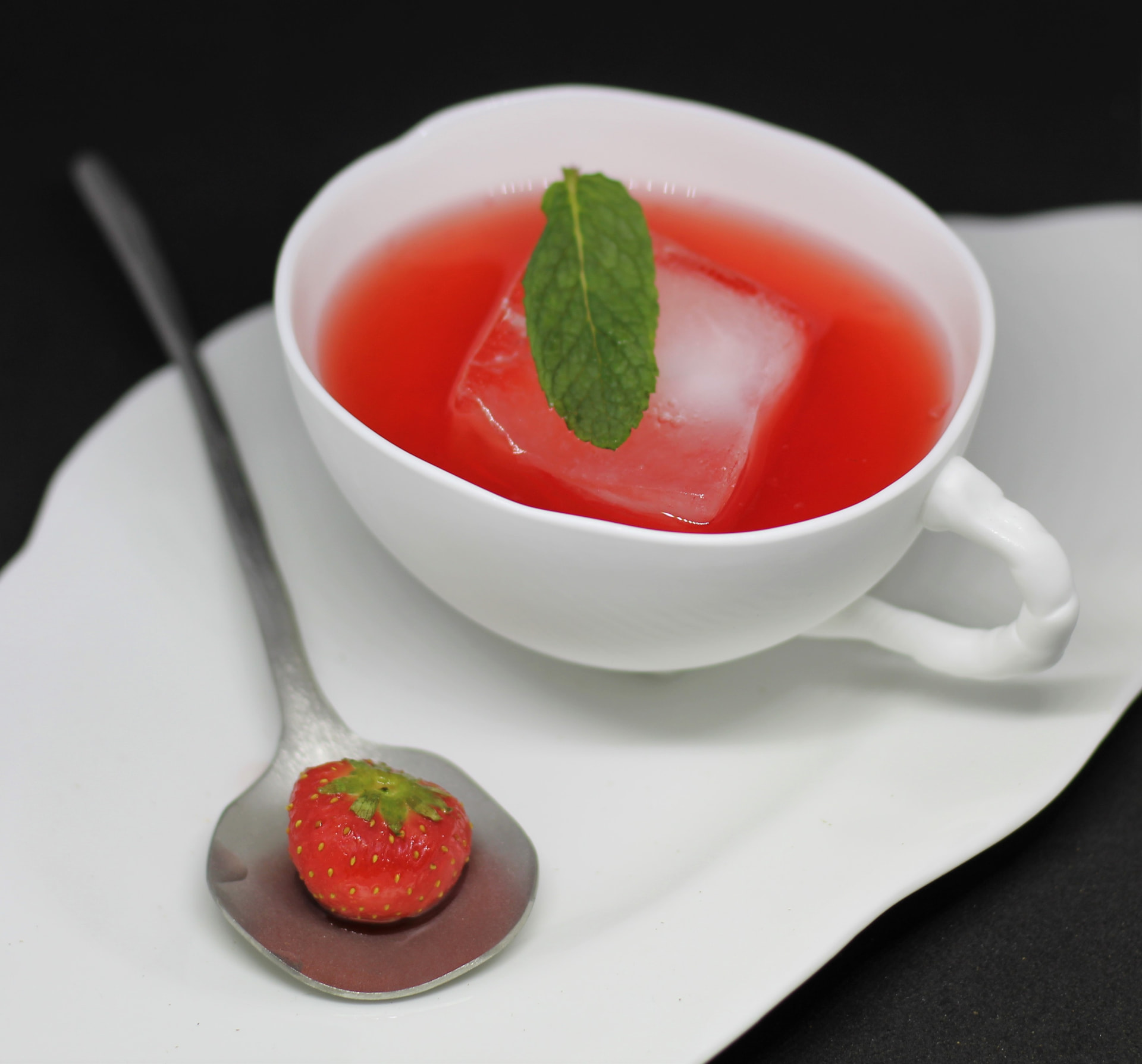 Red fruit serve in a porcelain cup with clear ice, mint and a strawberry on a spoon