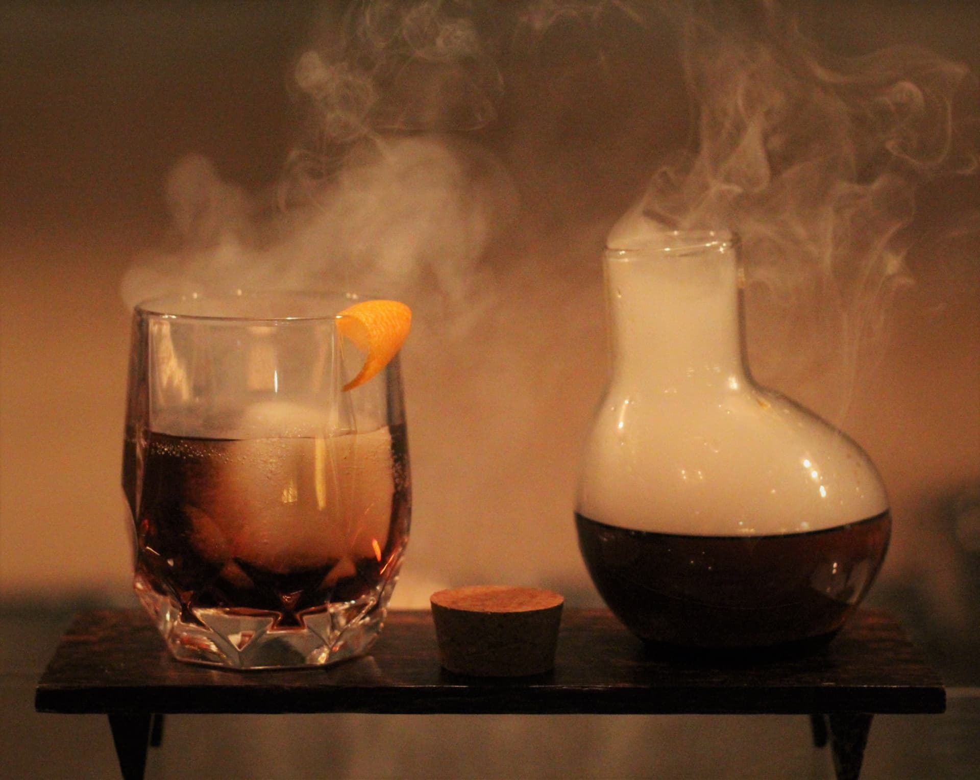 Smoked cocktail with orange twist beside a smoking decanter on a dark wooden stand