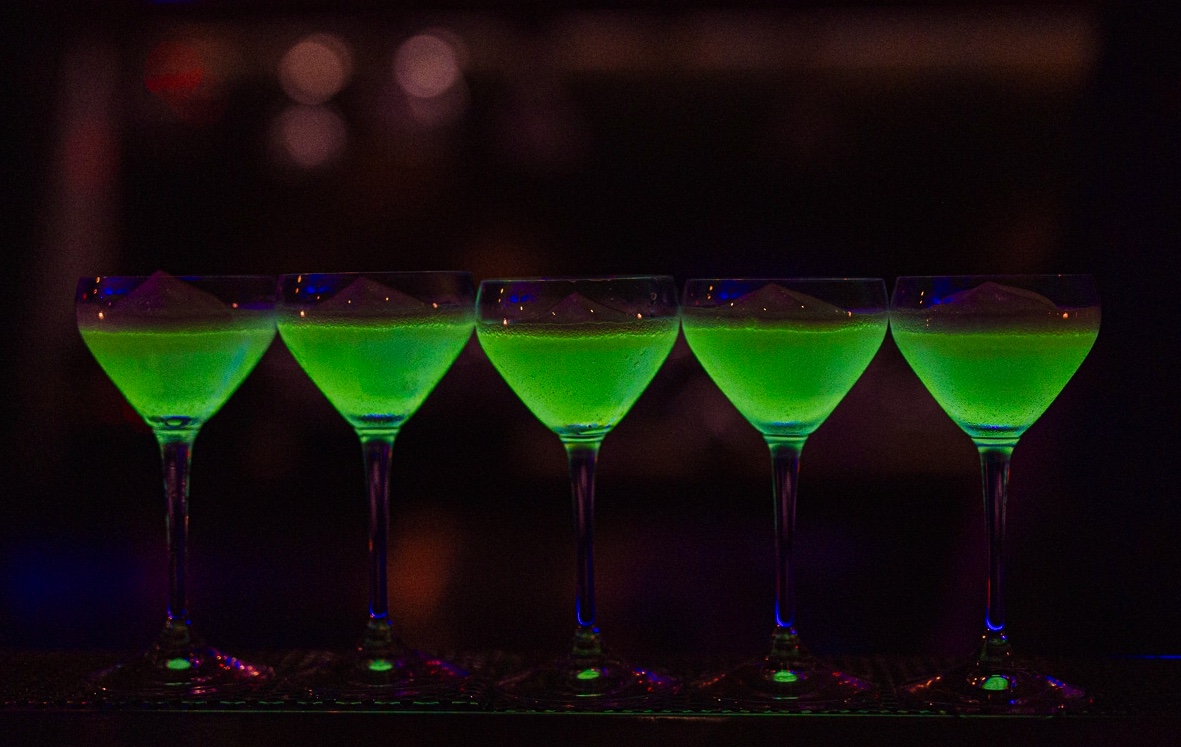 Row of five green cocktails lined up along the bar under blue light