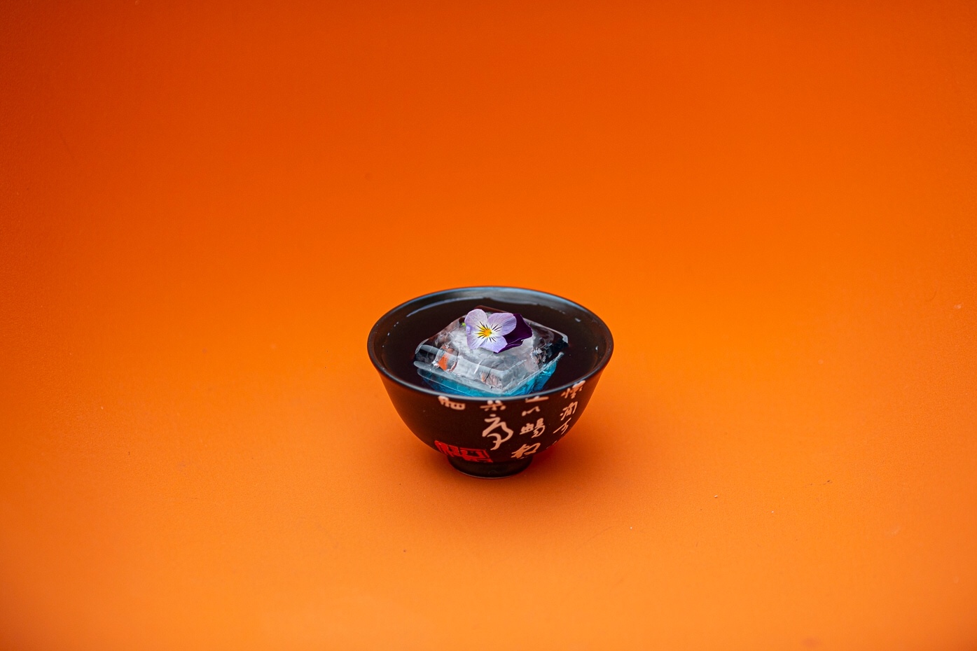 Blue serve in a small lacquered bowl with clear ice and a violet flower on orange