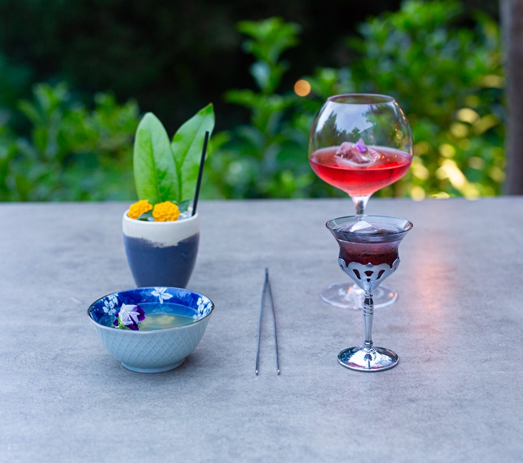 Four cocktails in mixed ceramic and crystal vessels on a garden table at dusk