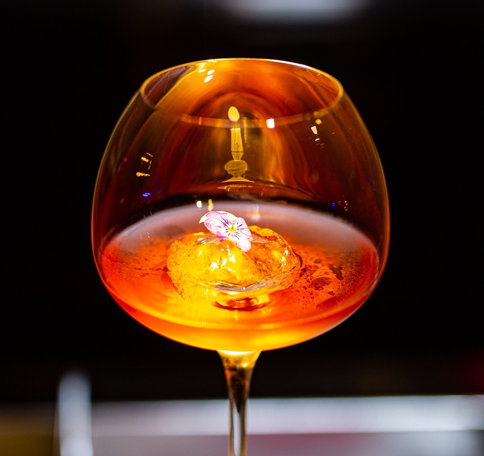 Backlit amber cocktail in a stemmed glass with a single violet flower