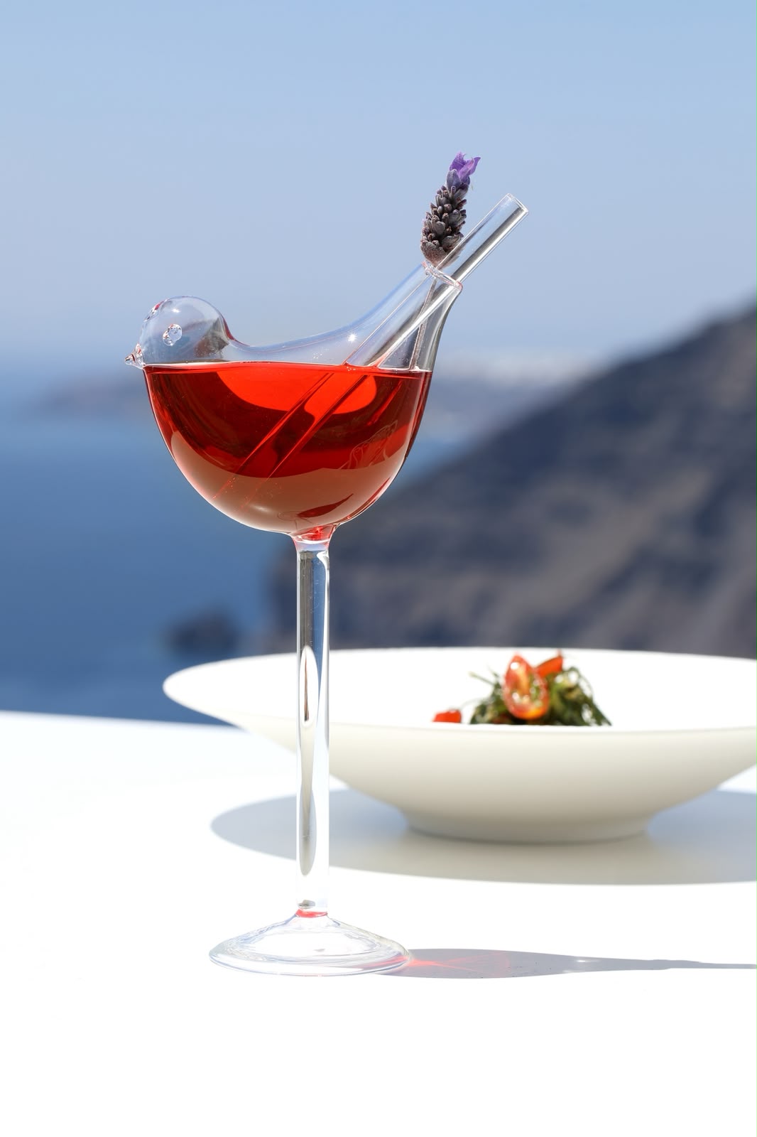 Bird-shaped glass with a red cocktail and lavender garnish above the Santorini caldera