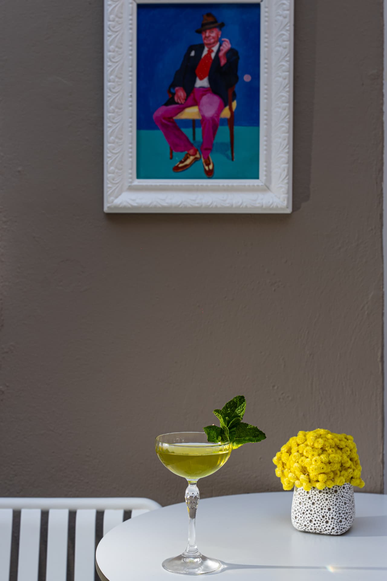 Green cocktail and yellow flowers on a white table below a framed painting