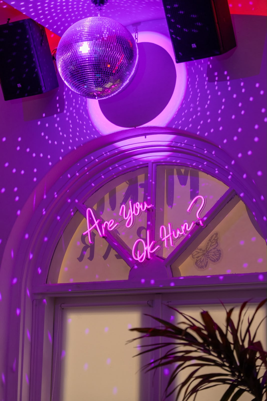 Disco ball and 'Are You OK Hun?' neon above the arched window in violet light