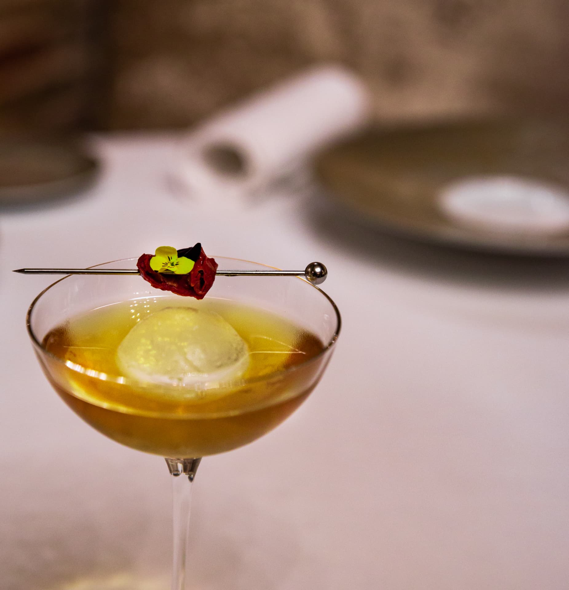 Close-up of an amber cocktail with a clear ice sphere and edible flower
