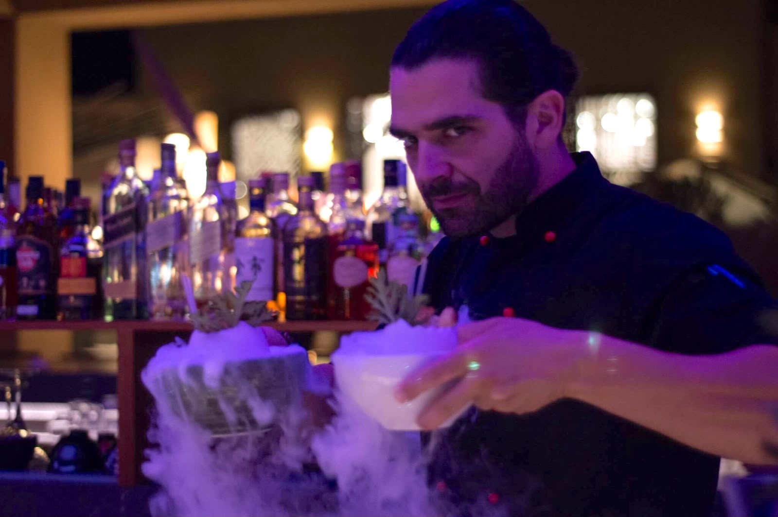 Bartender serving two smoking cocktails behind the lit back bar