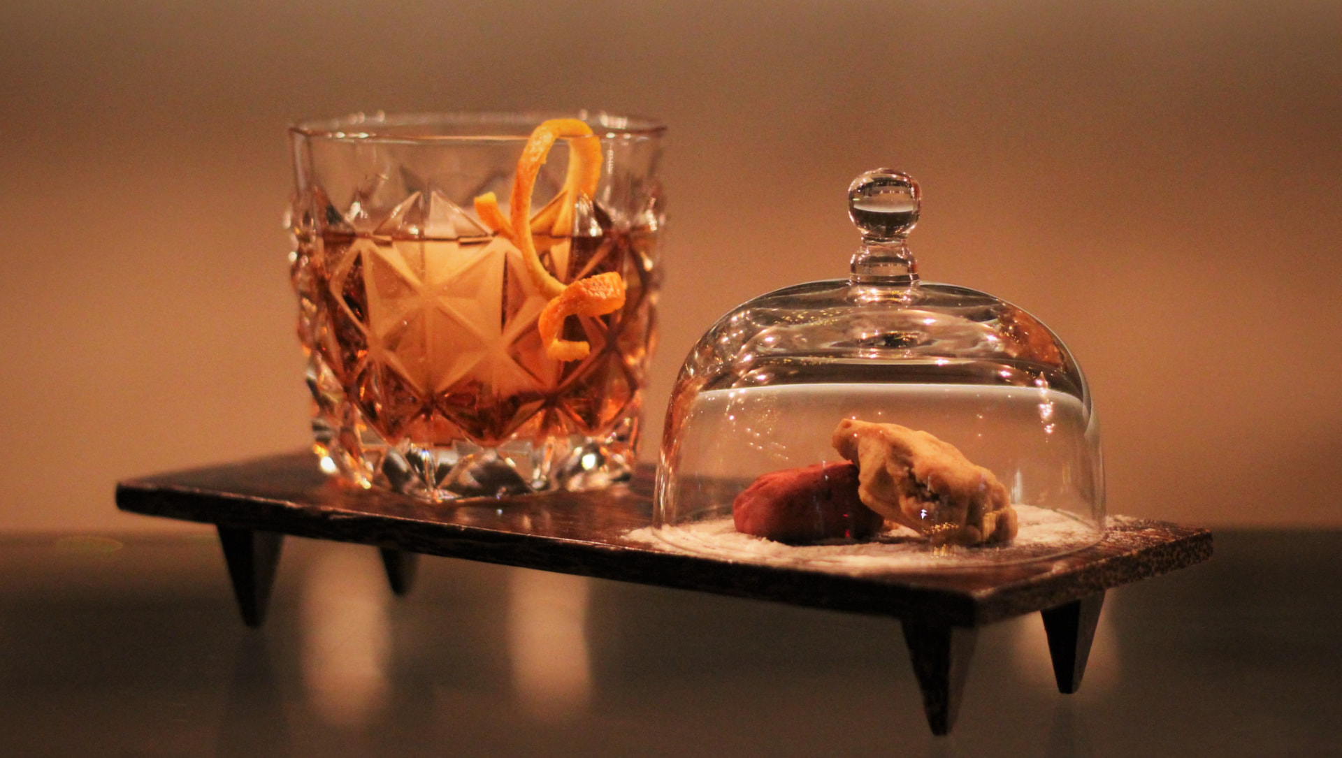 Cut-crystal old fashioned with orange twist beside a glass cloche and pastry