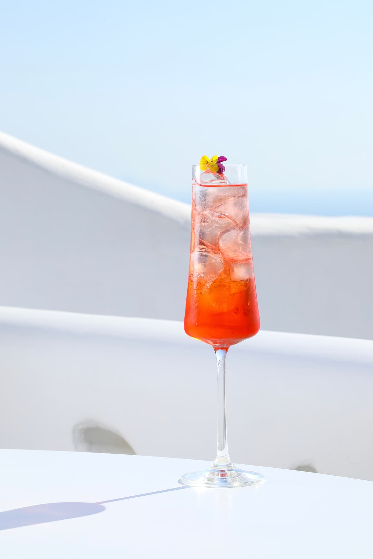 Tall spritz flute with edible flowers against white walls and sea light