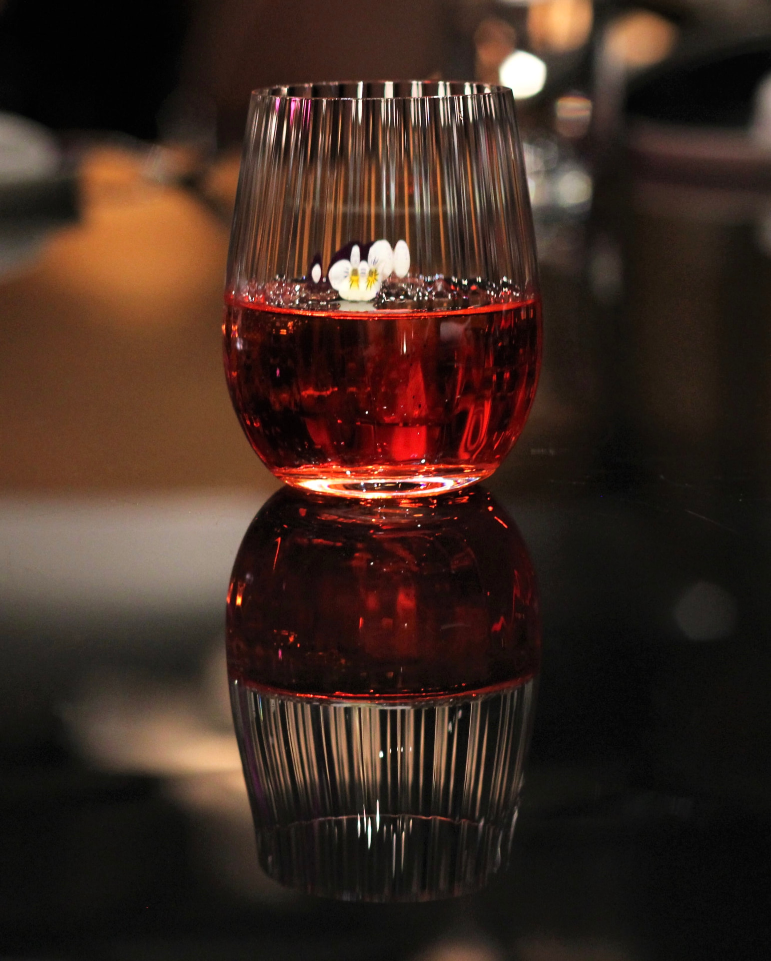 Ribbed tumbler of red aperitif with violet flowers, mirrored on the bar top